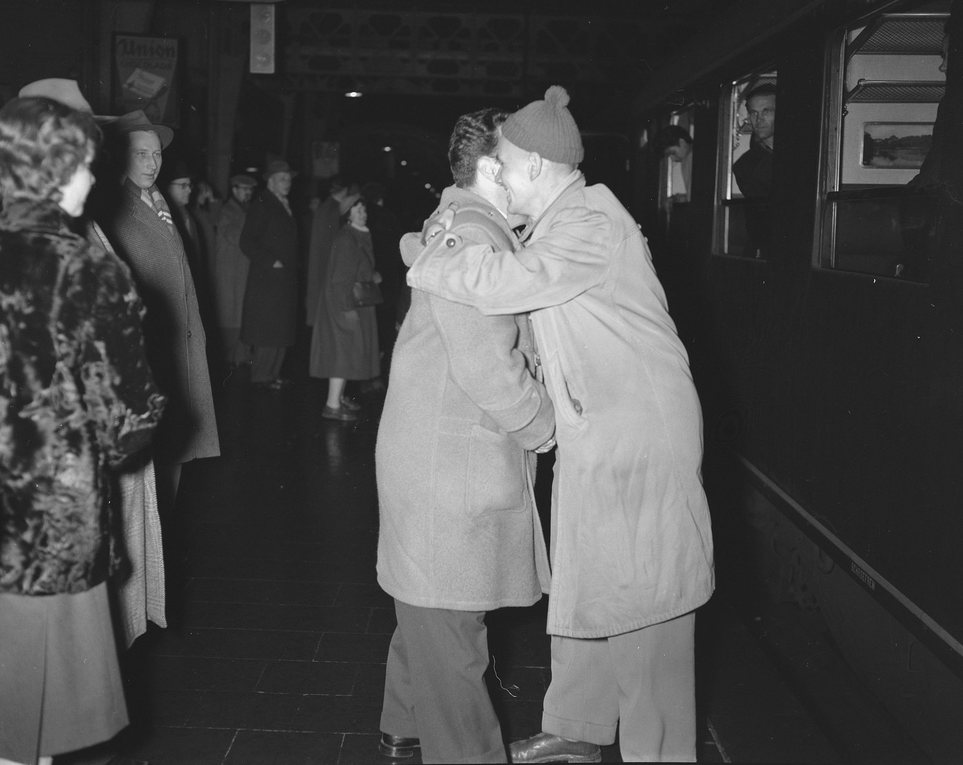 Two men in heavy coats, one in a knitted cap, embrace on a dim railway platform beside a train while women and men in winter coats look on