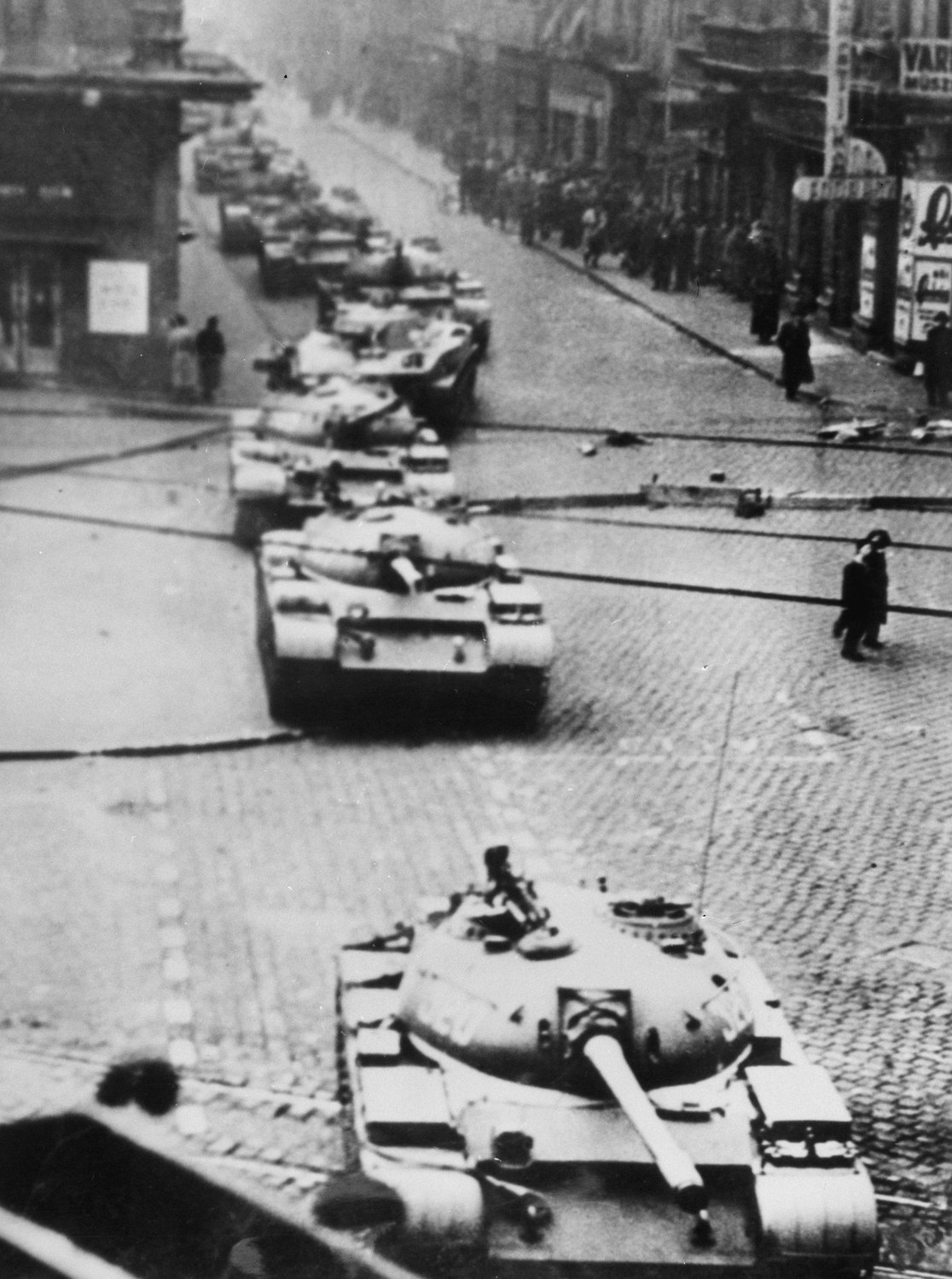 A column of tanks with long guns drives along a wide cobbled city street lined with shops and tall buildings, with a few pedestrians on the pavements