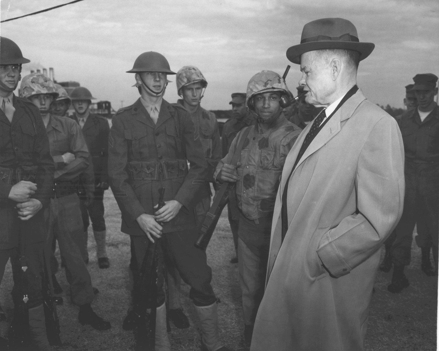 Black and white photograph of Puller in a hat and long coat facing a group of helmeted Marines