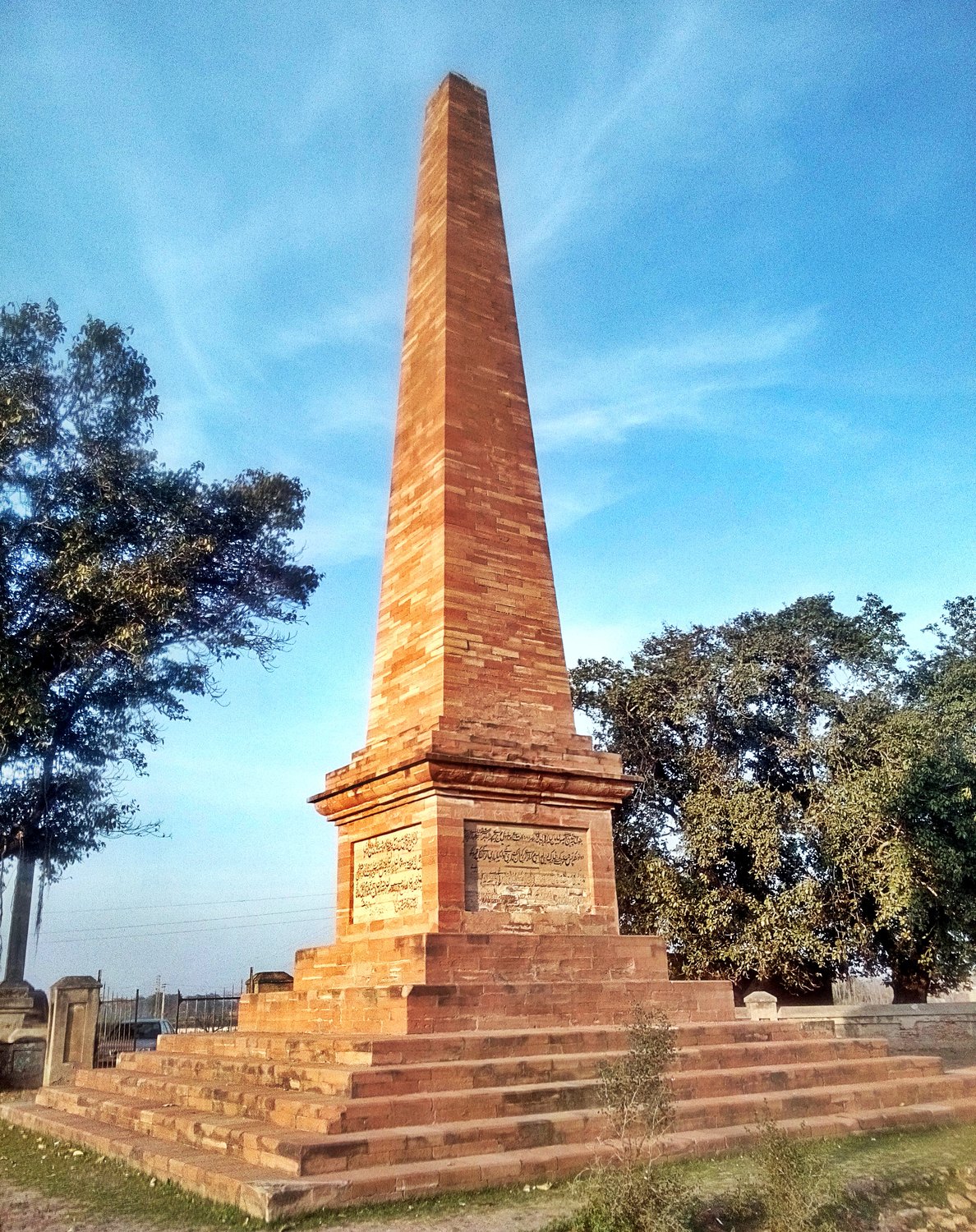 Tall tapering obelisk of red stone on a stepped base with two inscribed panels, standing among trees under a blue sky