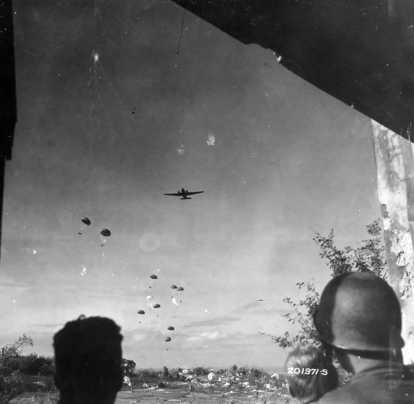 Seen past the silhouetted heads of two soldiers under a shattered concrete roof, a twin engine transport flies over Topside while a string of small cargo parachutes drifts down onto ground littered with canopies