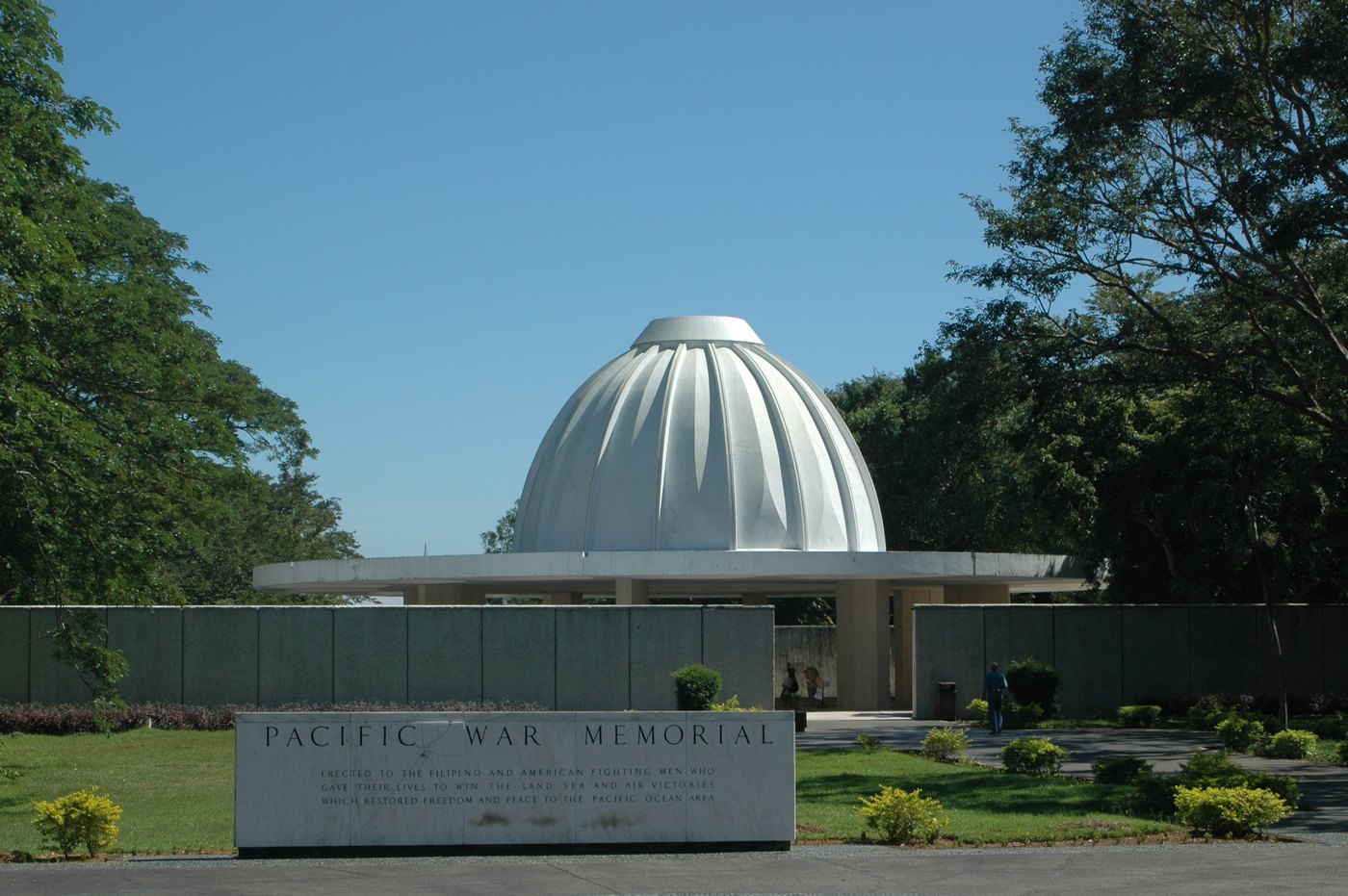 A white fluted dome on slender columns rises behind a low gray wall and a stone tablet reading Pacific War Memorial, among green lawns and tall trees under a clear blue sky