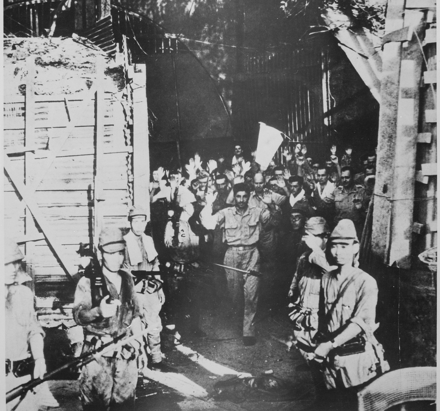 Black and white photograph of a crowd of American soldiers with raised hands and a white flag coming out of a timbered tunnel entrance, watched by Japanese soldiers with rifles and fixed bayonets in the foreground