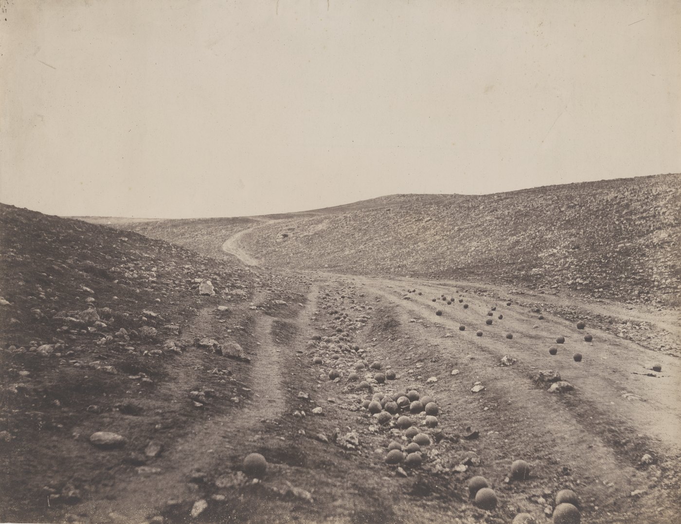 Sepia photograph of an empty, stony ravine with a dirt road strewn with cannonballs, under a pale sky.
