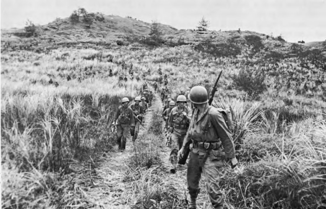 Black and white photograph of a file of American infantrymen walking along a grassy ridge