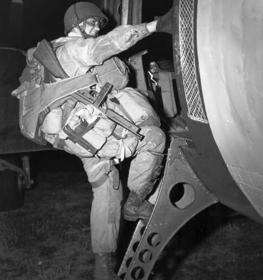 Black and white photograph of a heavily laden paratrooper climbing through the door of a transport aircraft at night