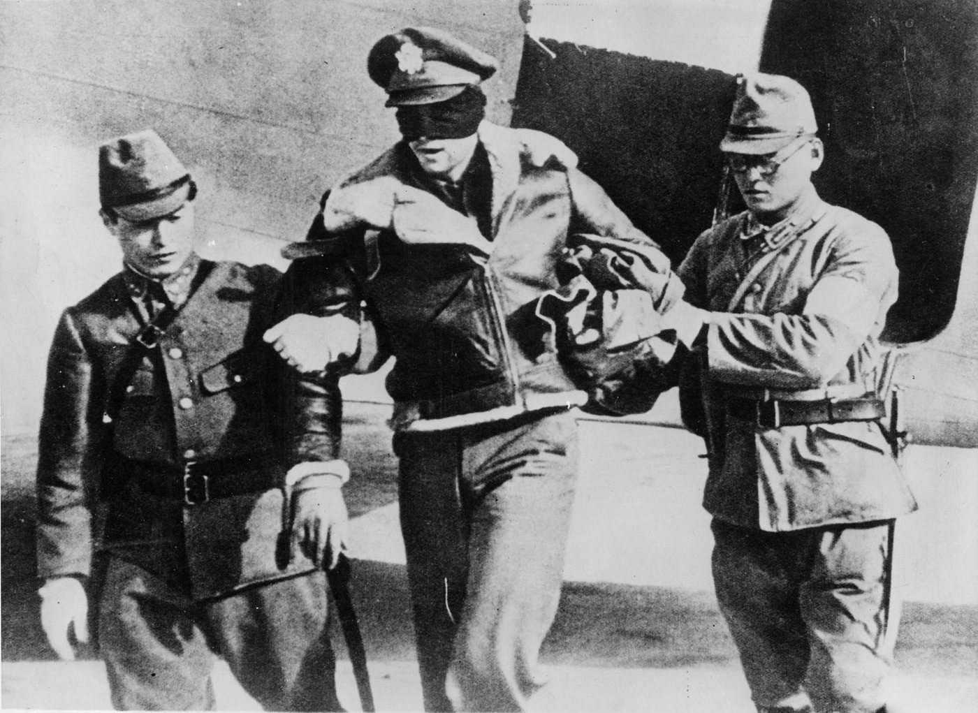 Black and white photograph of a blindfolded American airman in a flight jacket held by the arms by two Japanese soldiers