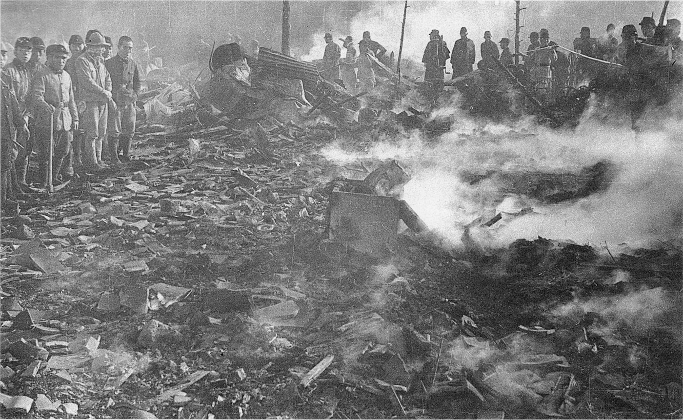 Black and white photograph of smoking rubble in a Tokyo street with civil defence men and police standing around it