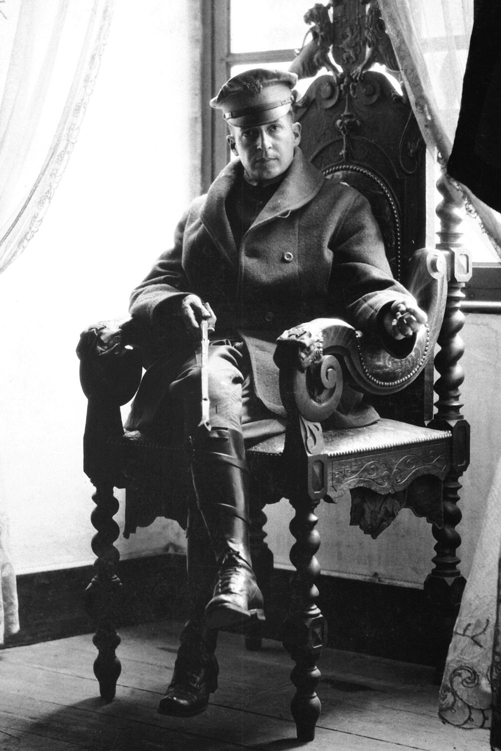 Black and white photograph of a young MacArthur in uniform and riding boots seated in an ornate chair