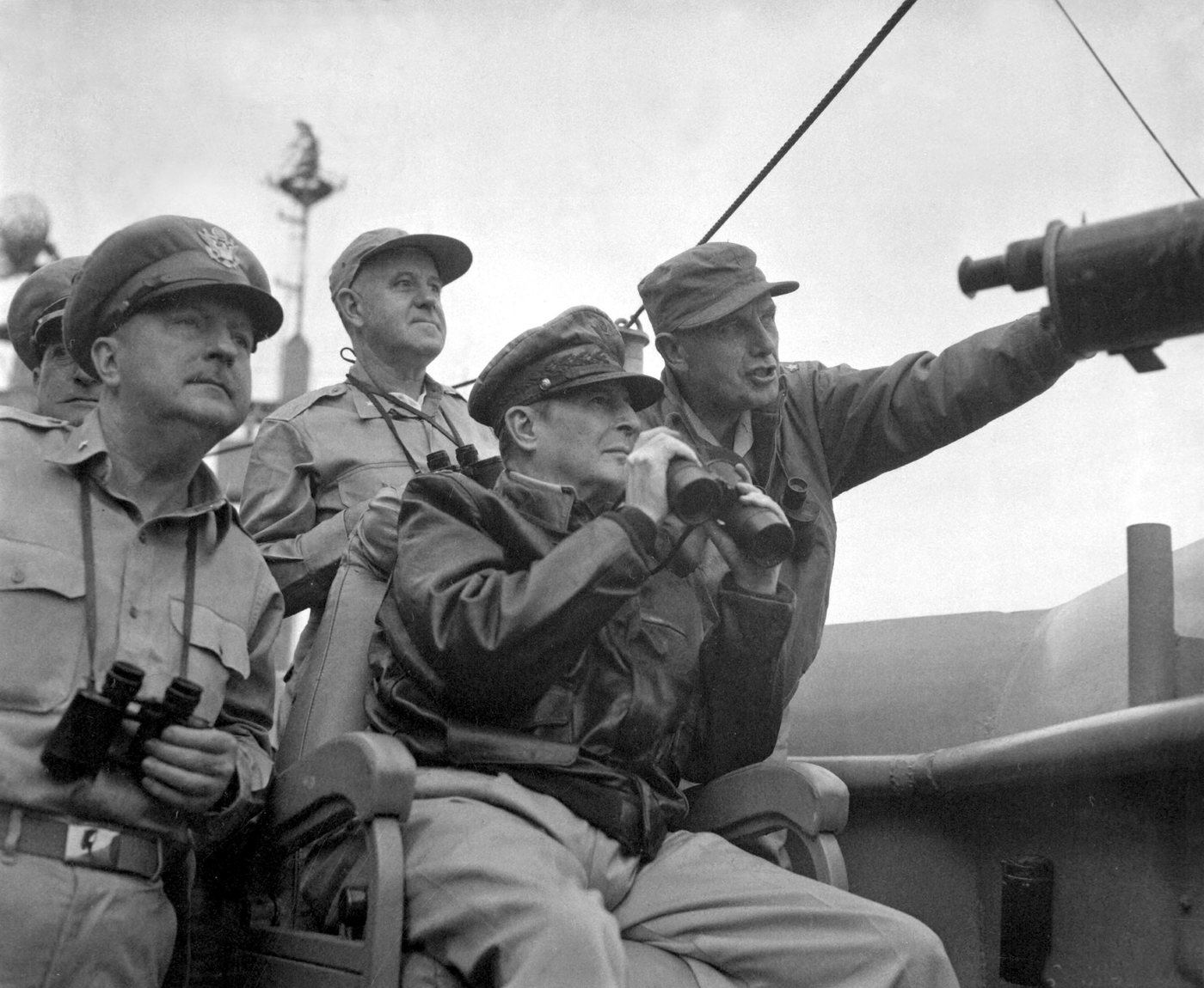 Black and white photograph of MacArthur in a peaked cap seated on a ship's bridge with officers standing around him