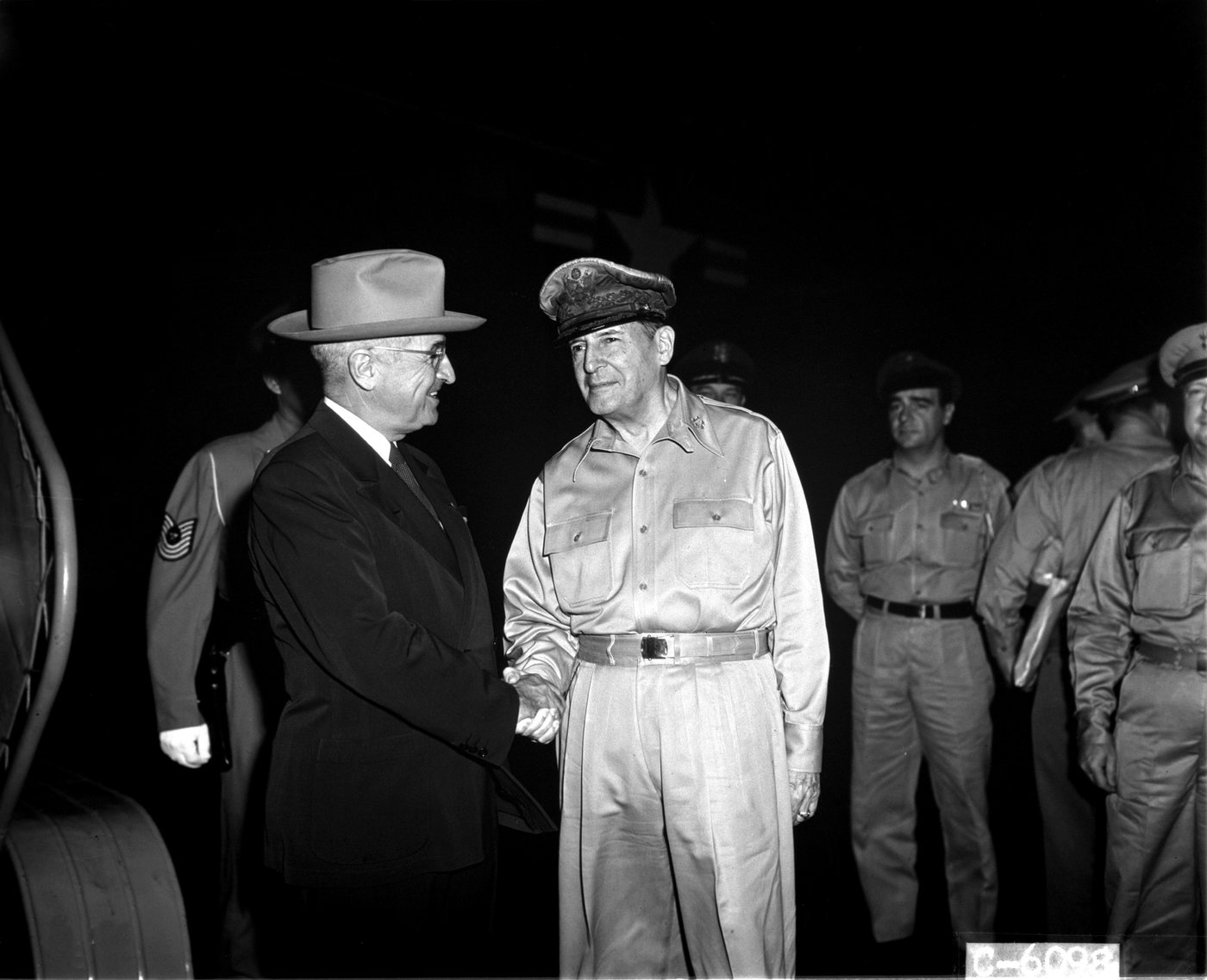 Black and white photograph of MacArthur shaking hands with President Truman beside an aircraft