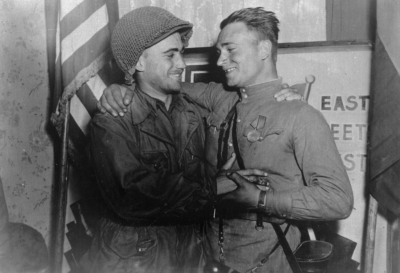 Black and white photograph of two young soldiers, an American in a netted helmet and a Soviet officer with a medal on his tunic, embracing and smiling in front of a sign reading East Meets West.