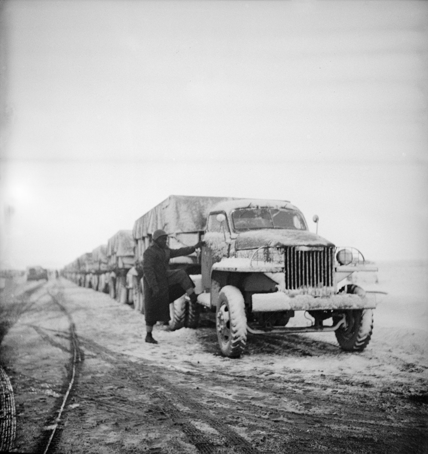 Black and white photograph: a long column of snow-covered, canvas-topped army trucks on a muddy road, with a soldier in a greatcoat climbing onto the running board of the first truck.
