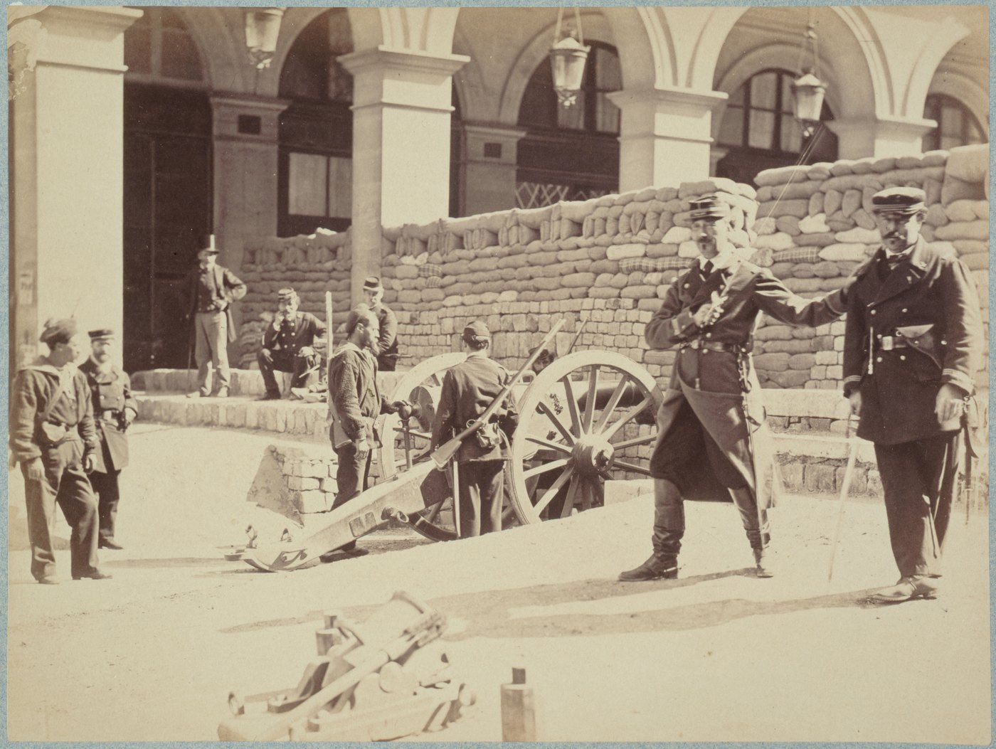 Sepia photograph of a street in Paris in 1871: National Guardsmen and civilians stand beside a cannon in front of a wall of sandbags and paving stones under an arcade.