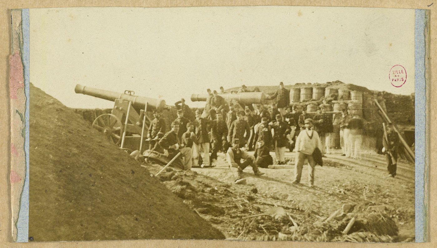 Faded sepia photograph of French soldiers and civilians posing around two heavy guns on an earthwork rampart, with barrels and gabions stacked behind them.