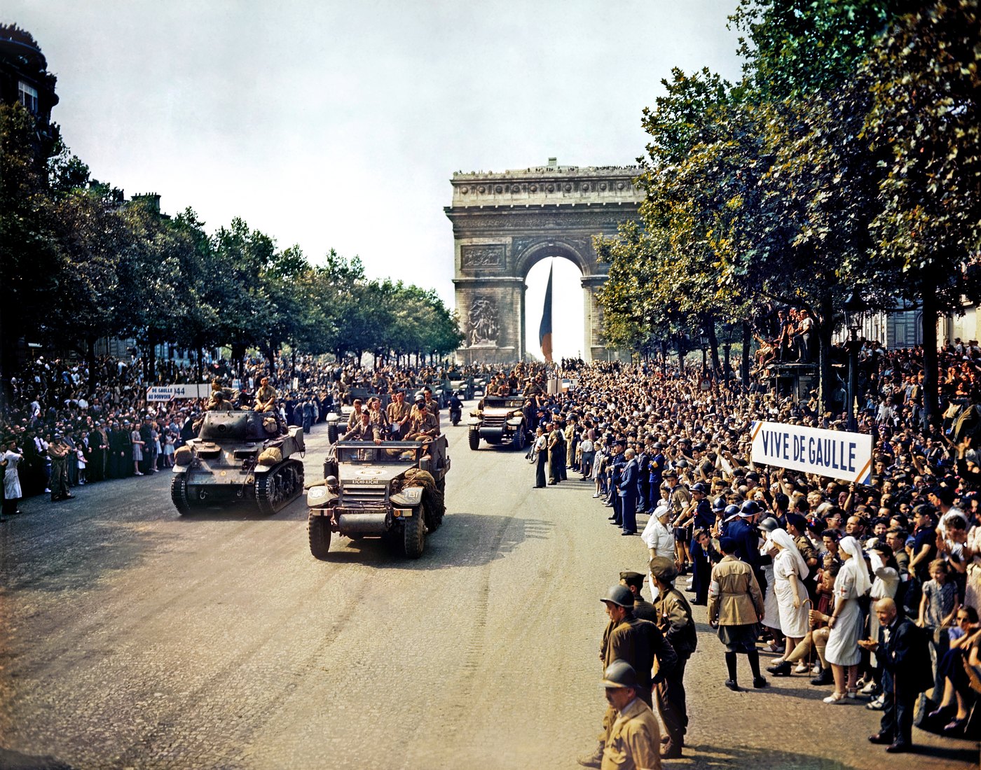 Color photograph of light tanks and half-tracks driving down the Champs-Élysées toward the Arc de Triomphe between dense crowds, one holding a Vive de Gaulle banner.