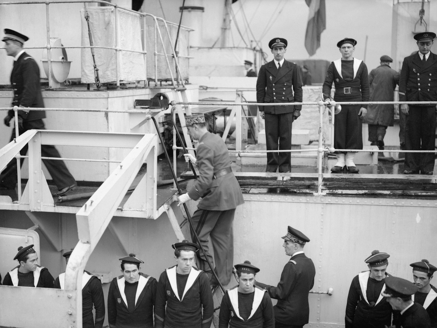 A general in a kepi and belted uniform climbs a ladder onto a ship's upper deck while French sailors with wide collars and officers in dark uniforms stand to attention