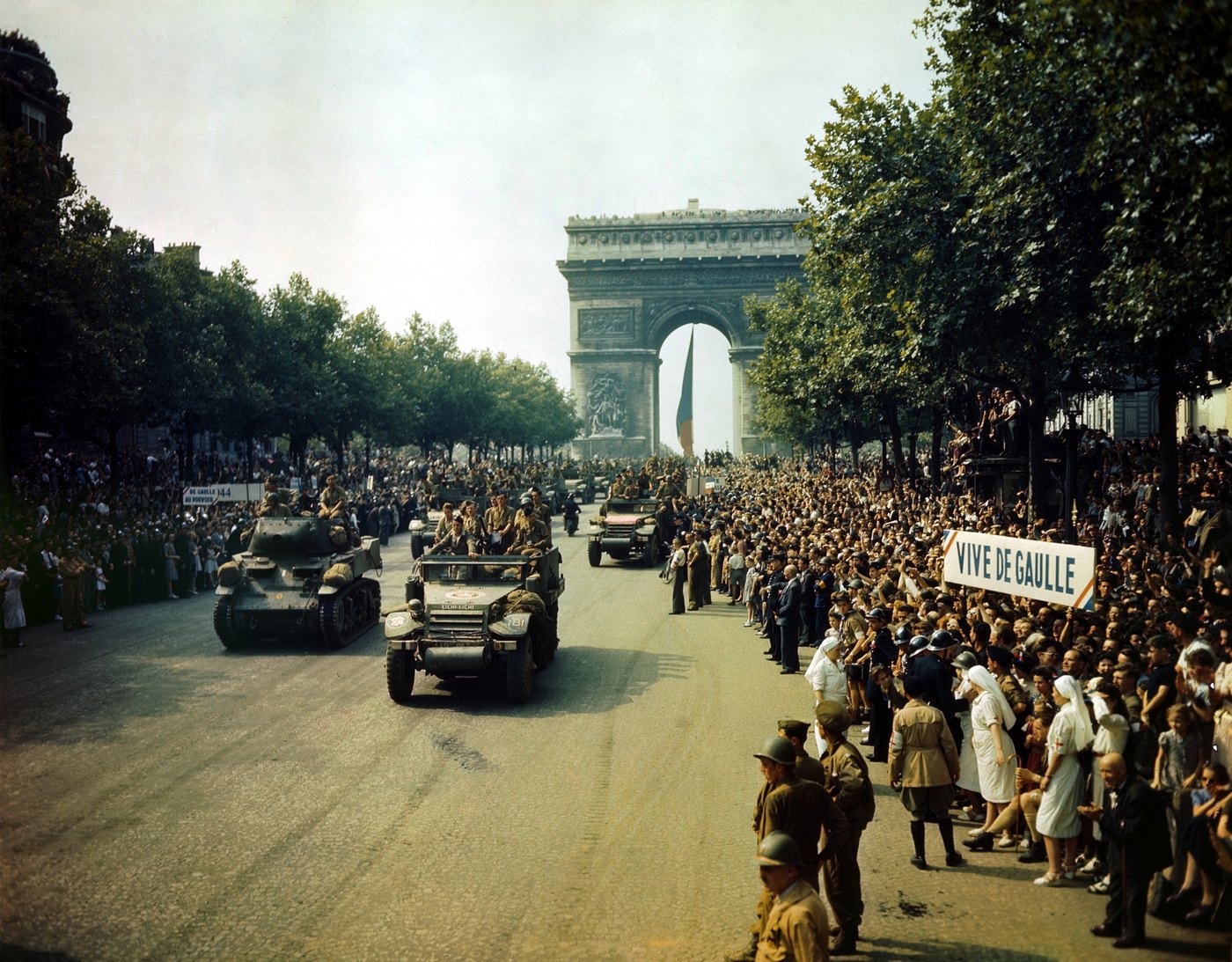 Color photograph of tanks and half tracks driving down a wide tree lined avenue from the Arc de Triomphe between dense crowds, with a banner reading Vive de Gaulle on the right