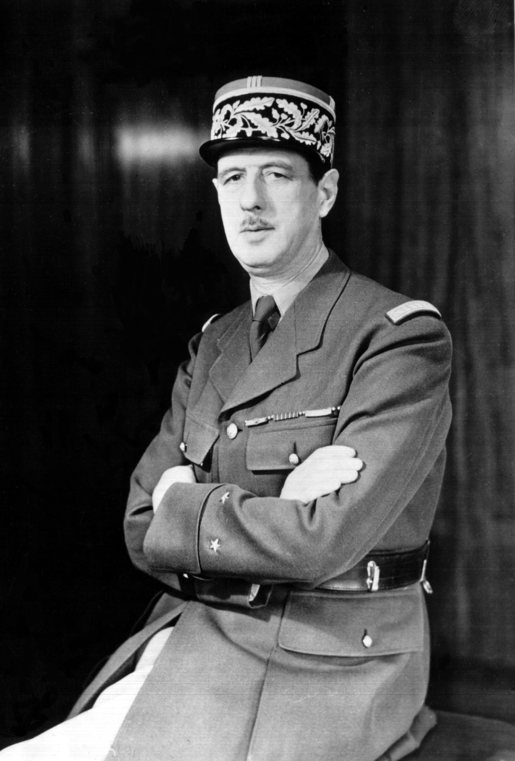Black and white studio portrait of a tall officer with a small mustache, arms folded, in a kepi with embroidered oak leaves and a belted uniform tunic with two stars on the cuff