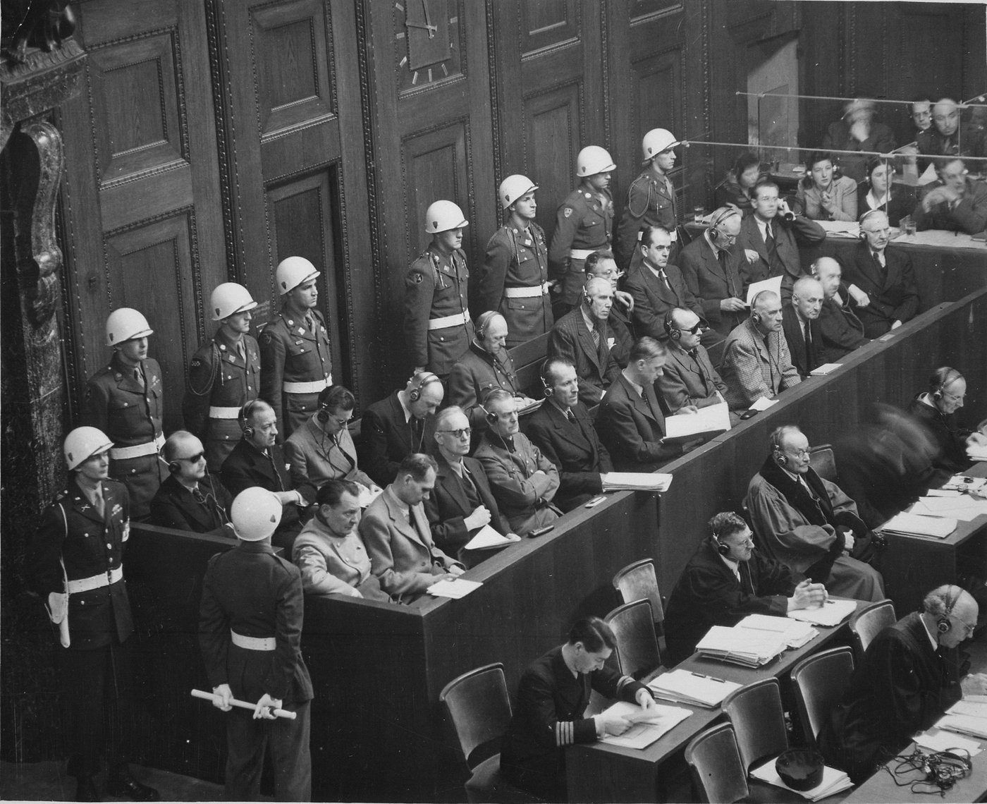 Black and white photograph of a courtroom from above: two rows of defendants wearing headphones sit in a wooden dock, guarded by military police in white helmets, with defense lawyers at desks in front.