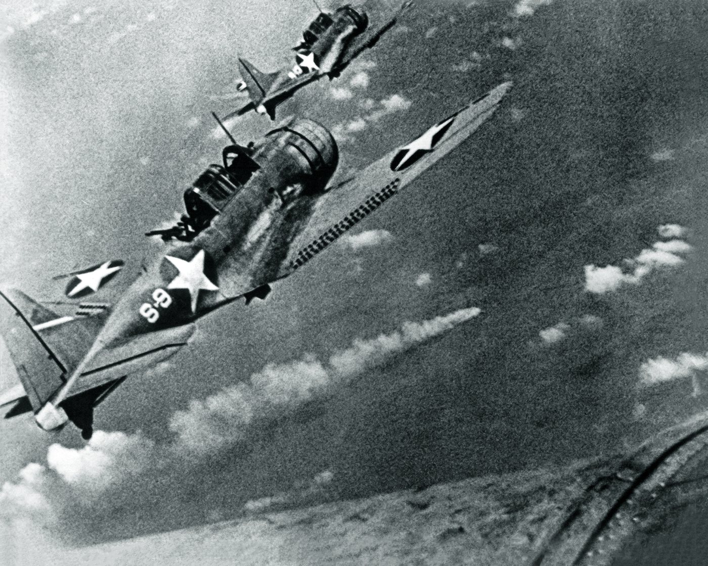 Two American dive bombers flying over a burning warship far below