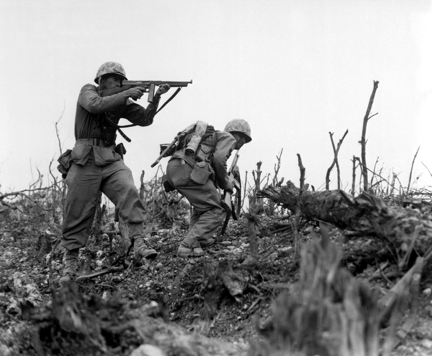 Marine aiming a submachine gun across shattered ground while a comrade ducks