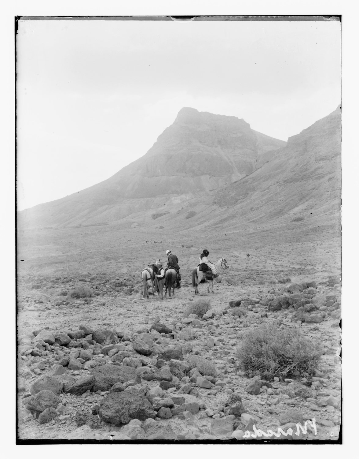 Black and white photograph of riders on stony ground below a steep desert mountain