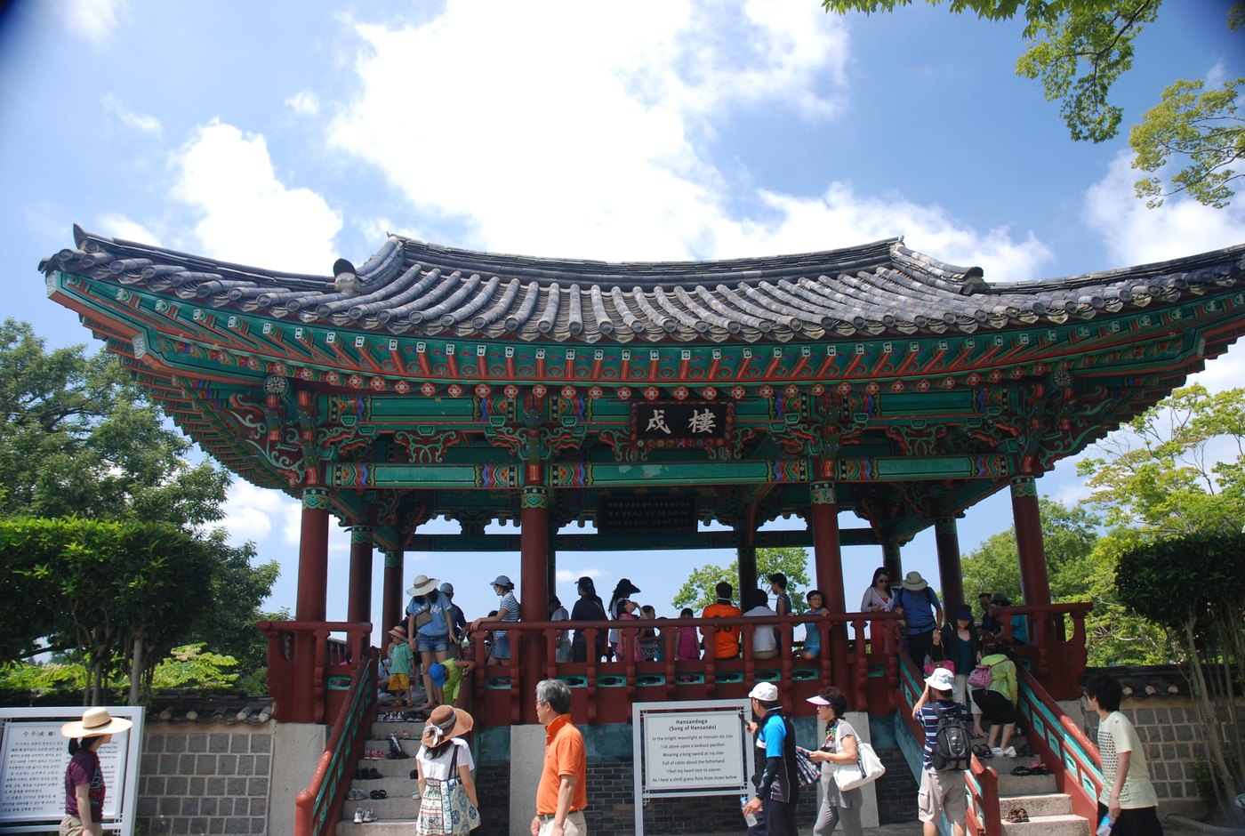 Visitors on the steps and floor of a painted wooden pavilion with a curved tiled roof, a signboard reading Hansandoga in front