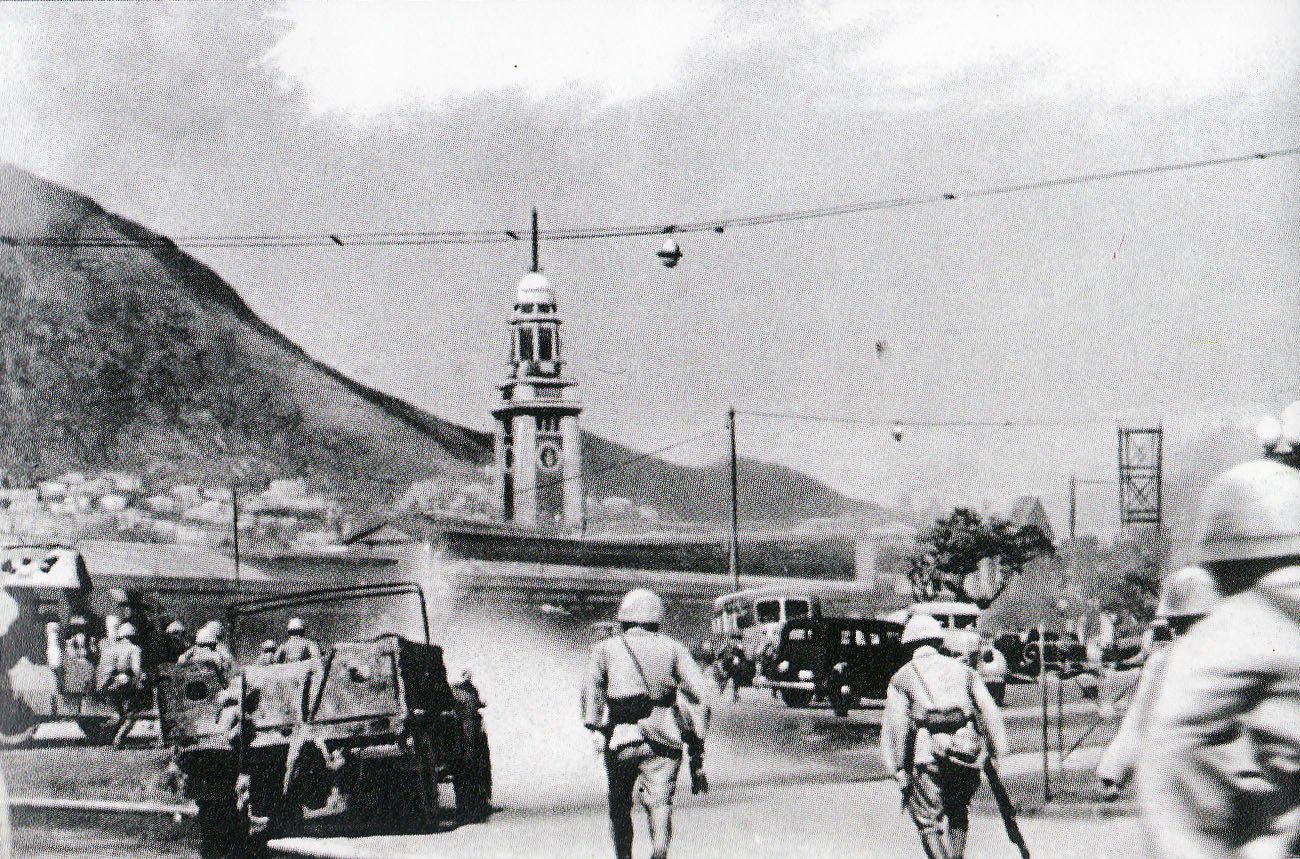Soldiers in helmets advance along a road towards a tall clock tower by the harbor, past abandoned cars and a bus, with dust in the air and steep hills behind