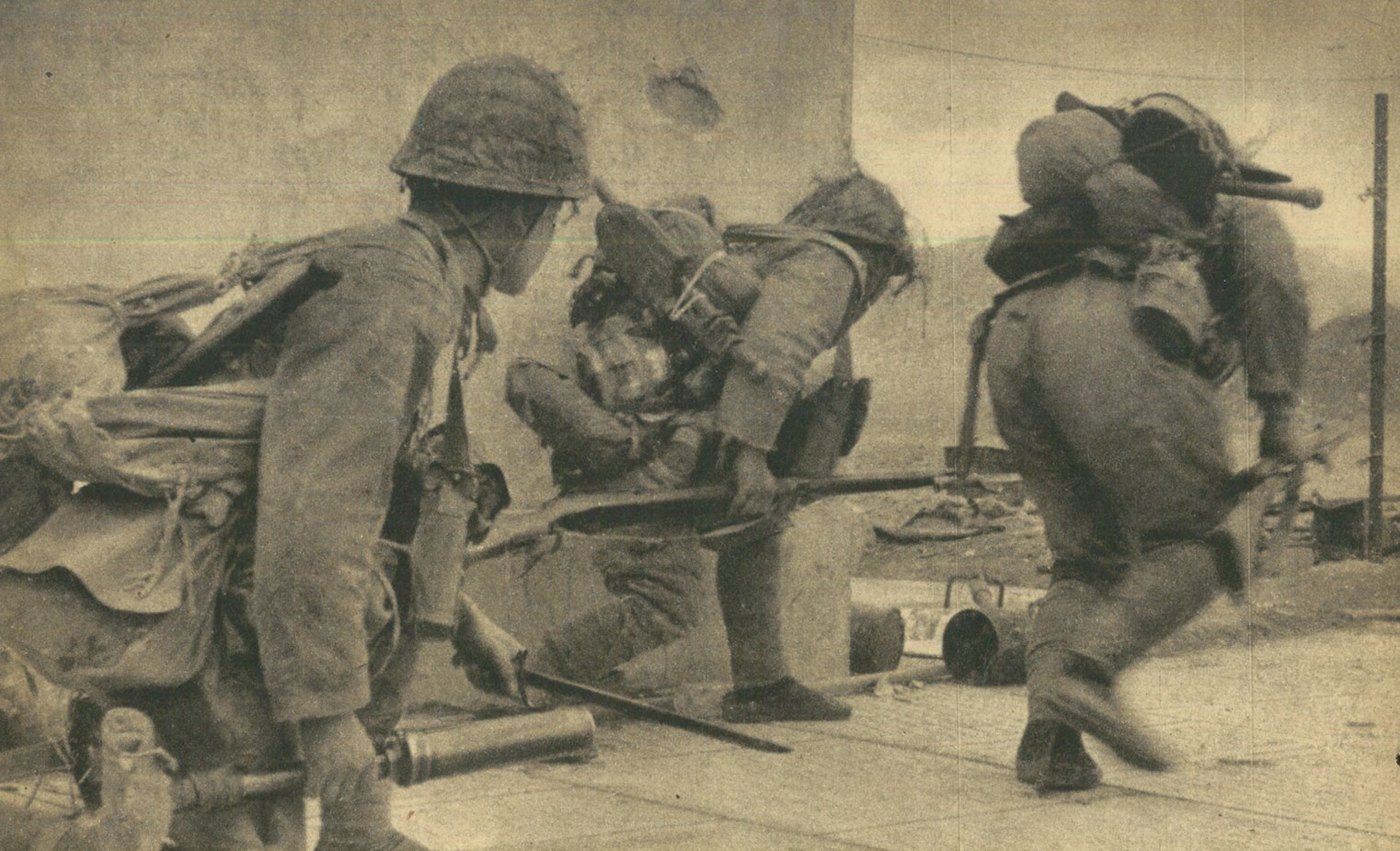 Three Japanese soldiers in netted helmets and heavy packs, rifles with fixed bayonets, run crouching past the corner of a pockmarked wall onto a paved street