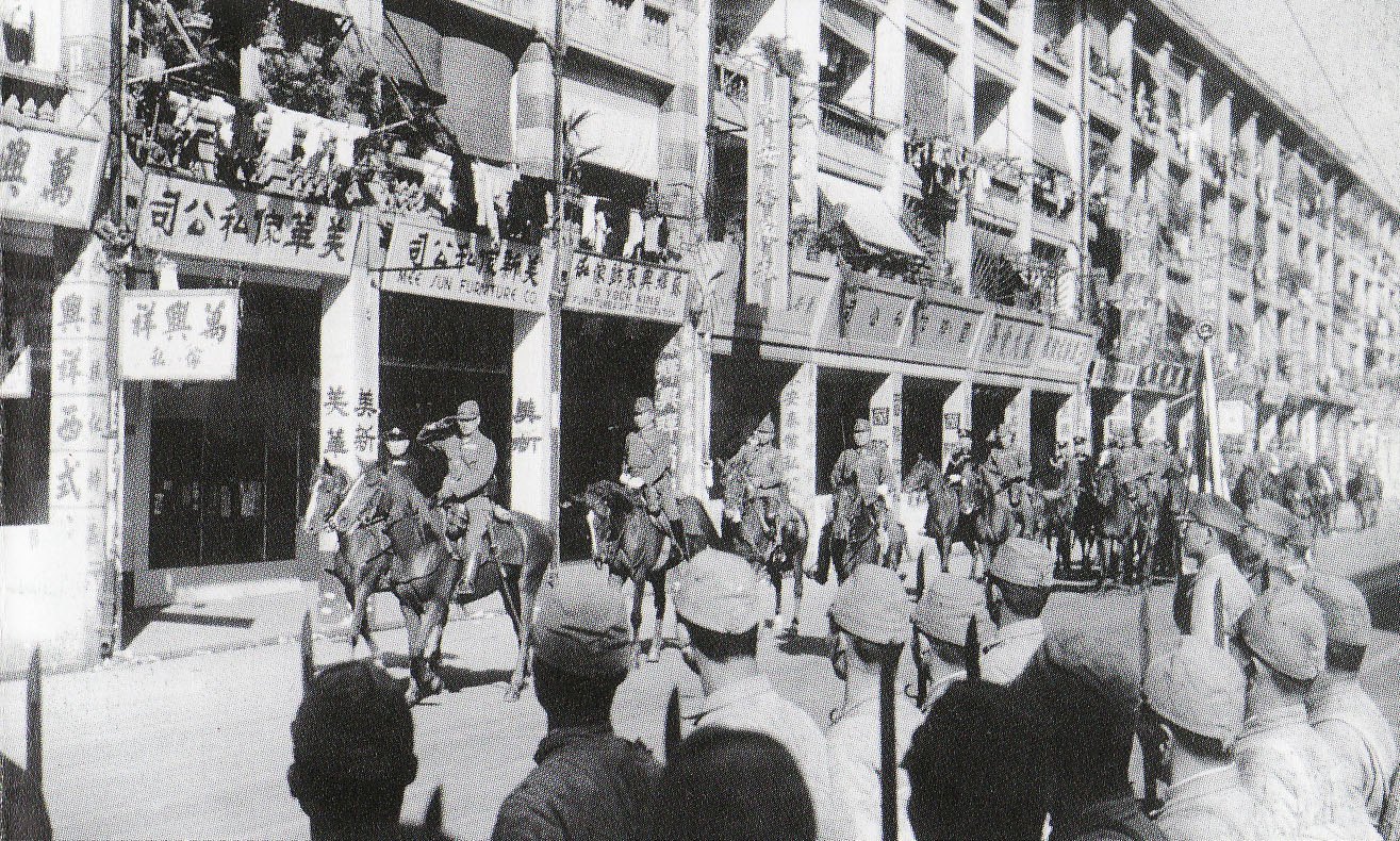Mounted Japanese officers, one saluting, ride past shophouses hung with Chinese signboards and laundry while rows of soldiers in caps stand in the foreground