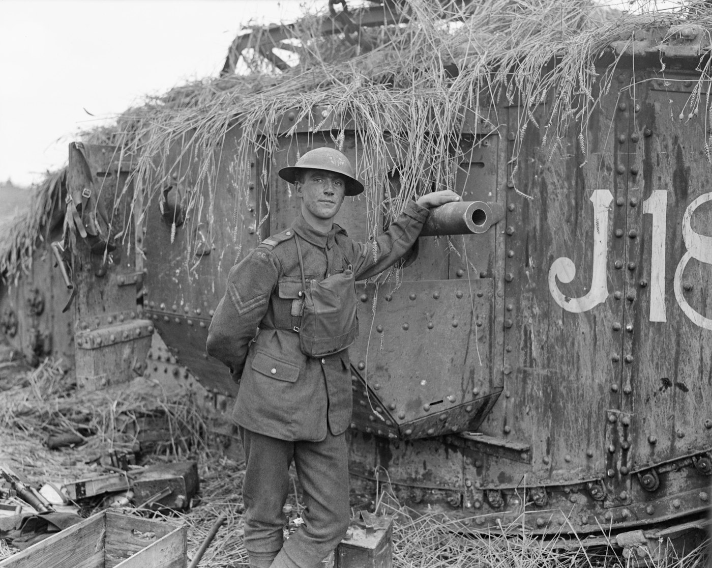 A young corporal in a steel helmet stands with one hand on the gun barrel of a heavy tank whose roof is covered in cut wheat as camouflage, the letters J18 painted on its riveted side