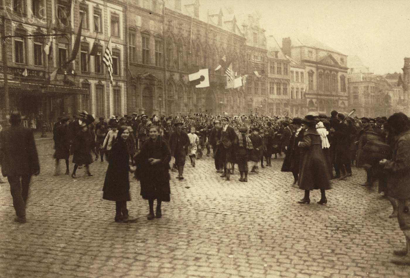 Sepia photograph of a crowded cobbled square lined with tall old buildings hung with flags, where pipers and soldiers march through civilians and two smiling girls walk towards the camera