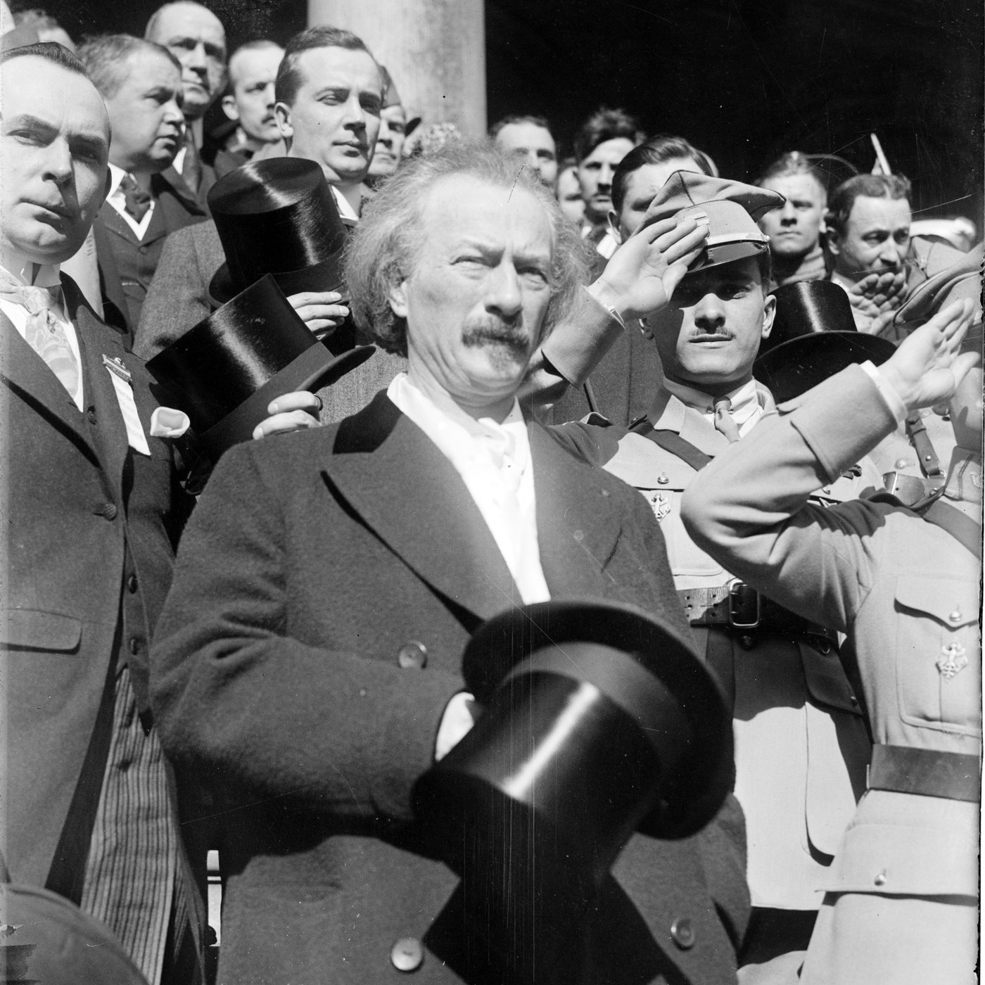 Paderewski z odkrytą głową, z cylindrem w dłoni, stoi w tłumie mężczyzn w cywilnych płaszczach, a obok niego salutują oficerowie w mundurach
