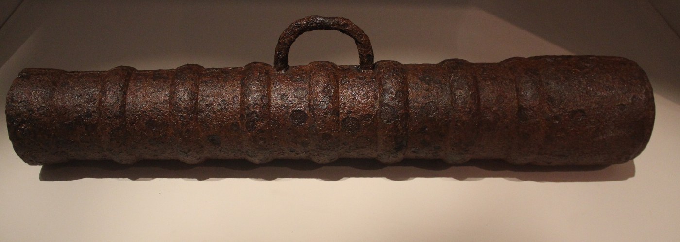 Heavily rusted iron cannon barrel lying horizontally in a museum display case, ringed with raised reinforcing bands, with a carrying handle on top.