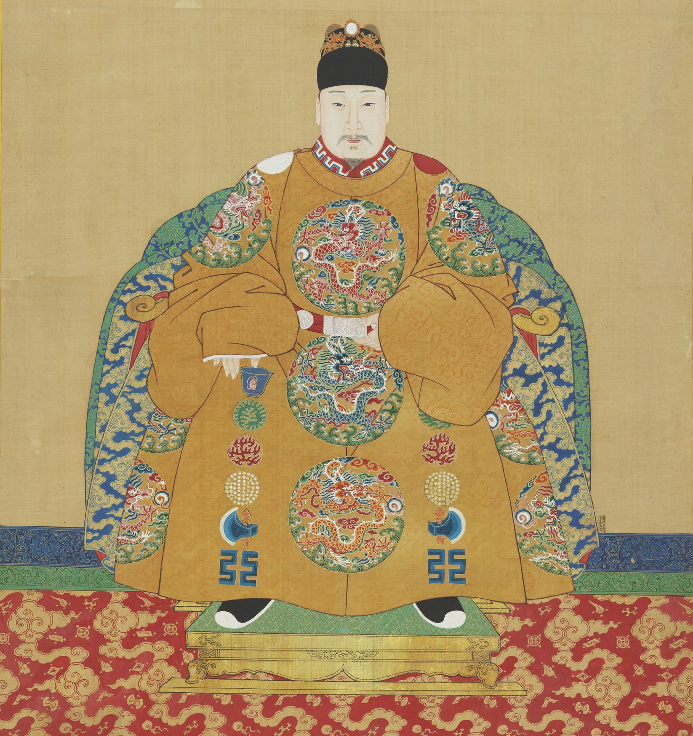Ming court portrait on silk of a seated emperor in a yellow robe embroidered with roundels of dragons, wearing a black cap with a gold ornament, his feet on a gilded footstool, above a red carpet patterned with gold clouds.