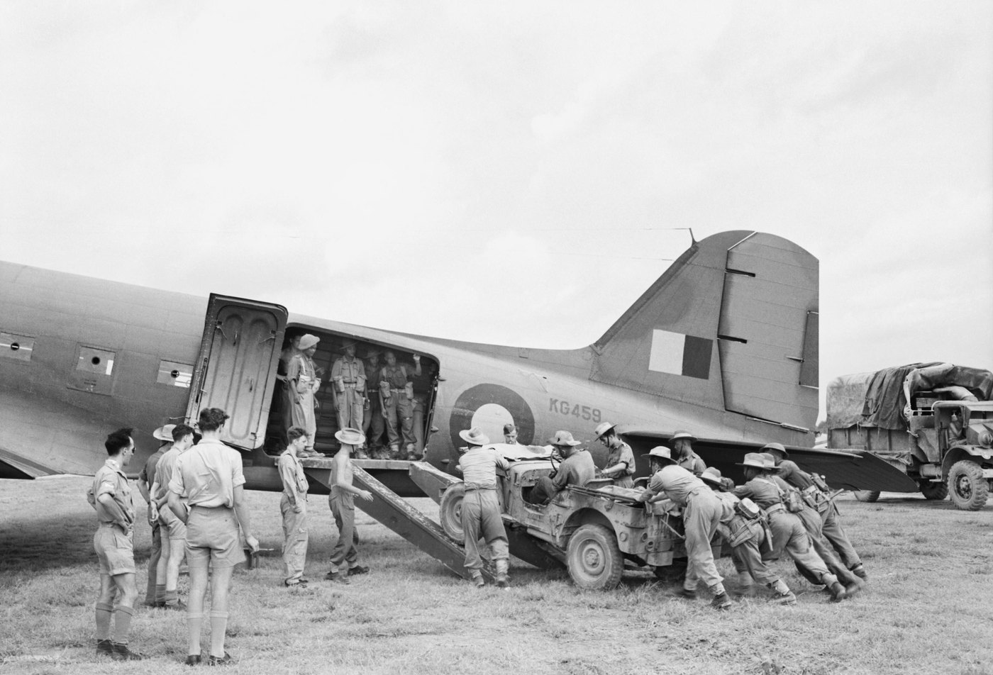 Soldiers push a muddy jeep up wooden ramps into the open cargo door of a twin engine transport aircraft marked KG459, while airmen and other troops watch from the grass