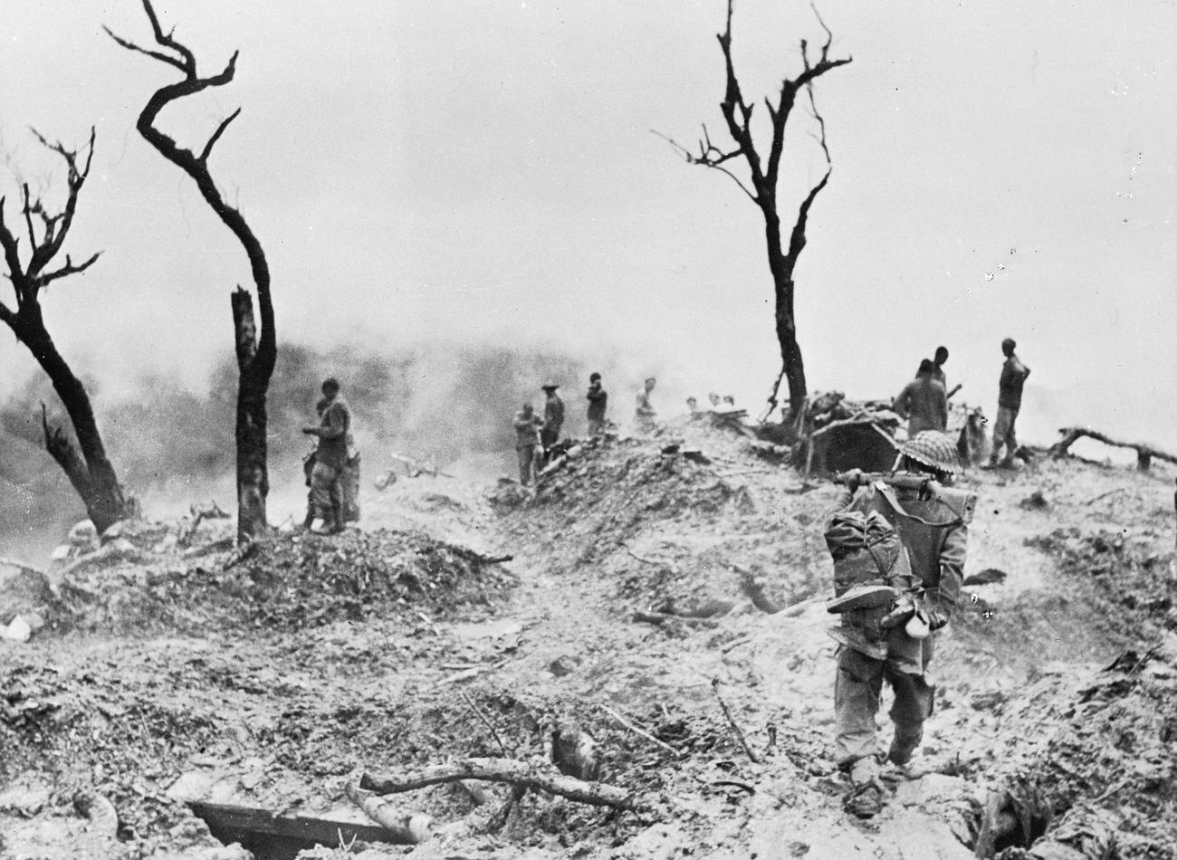 A soldier with a pack walks across a shell torn hilltop of mud, caved in bunkers and leafless shattered trees, with other soldiers standing in drifting smoke