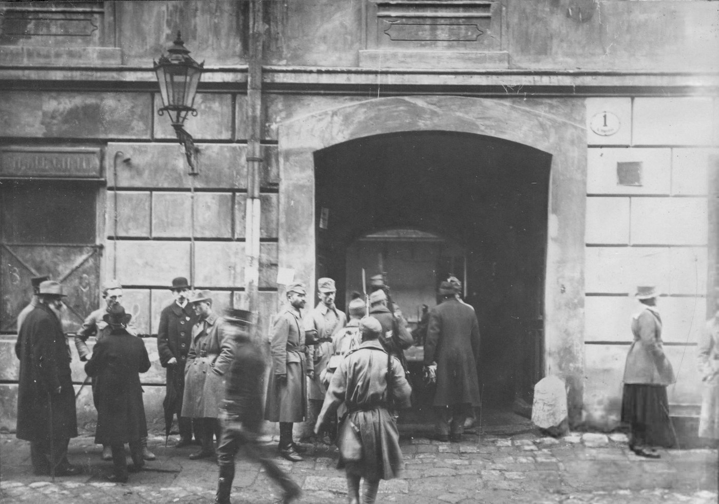 Black and white photograph of soldiers in caps and greatcoats, one with a rifle and fixed bayonet, and civilians in hats gathered at the arched entrance of a stone building numbered 1, with a street lamp on the wall and a woman standing at the right.