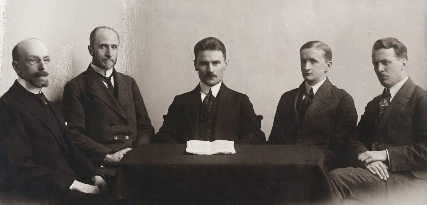Sepia studio photograph of five men in dark suits seated around a table covered with a dark cloth, an open booklet lying on the table in front of the man in the middle.