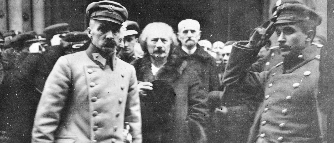 Black and white photograph of Józef Piłsudski in a peaked cap and a light double-breasted greatcoat walking past a crowd, followed by a white-haired man in a fur coat and an older man with a white moustache, while a young officer at the right salutes.