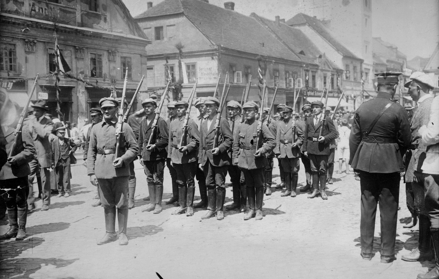 Black and white photograph of young men in flat caps and civilian jackets standing in ranks with rifles at their shoulders on a town square, with onlookers along the houses and a man in a peaked cap and belted uniform seen from behind at the right.