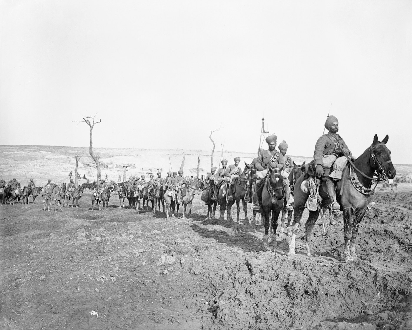 Black and white photograph of a long column of turbaned cavalrymen with lances riding in single file across a churned, muddy landscape with shattered tree trunks, the leading rider close to the camera on a dark horse.