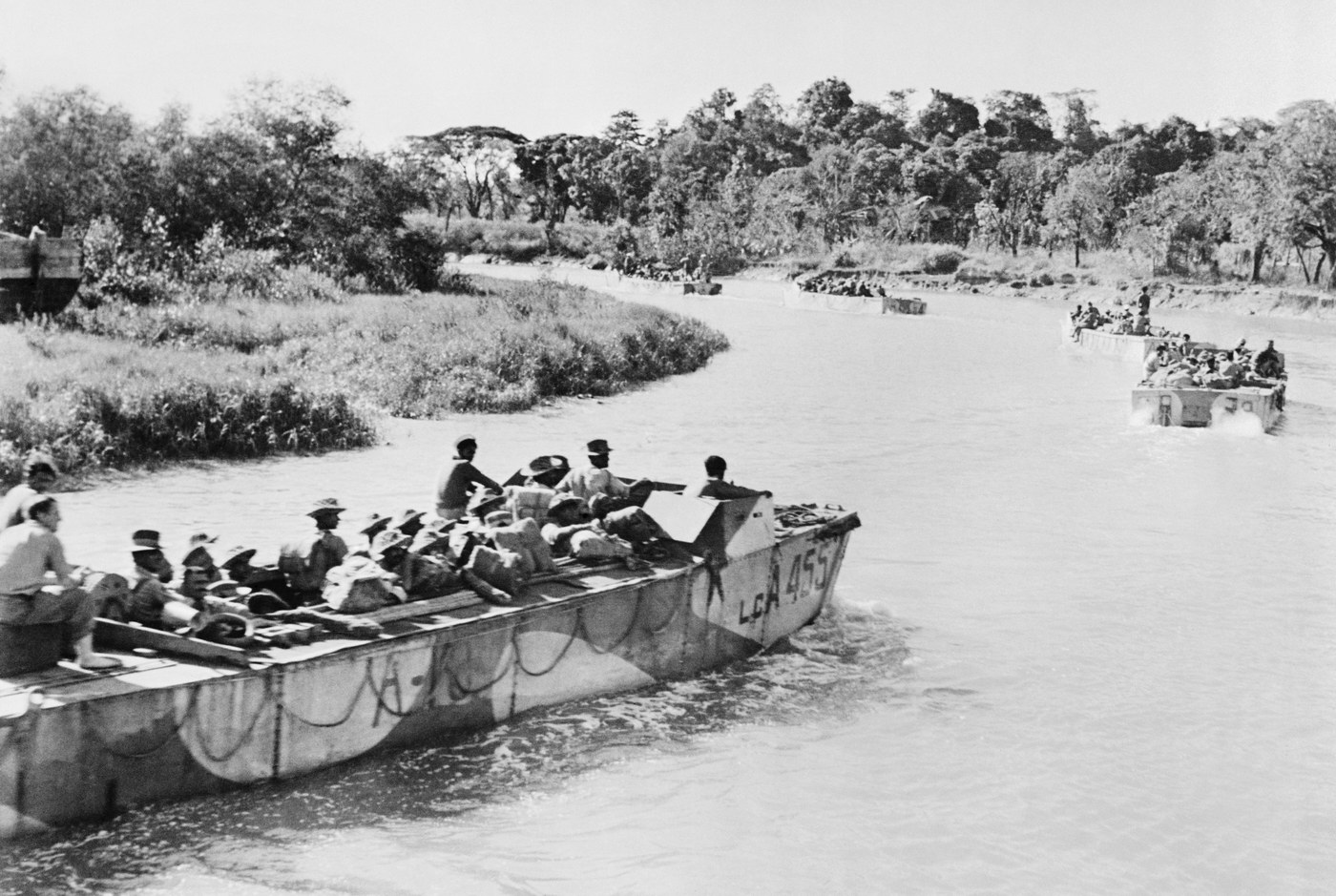 Black and white photograph of camouflaged landing craft full of soldiers and supplies moving in a line along a winding jungle river, with thick trees on both banks.