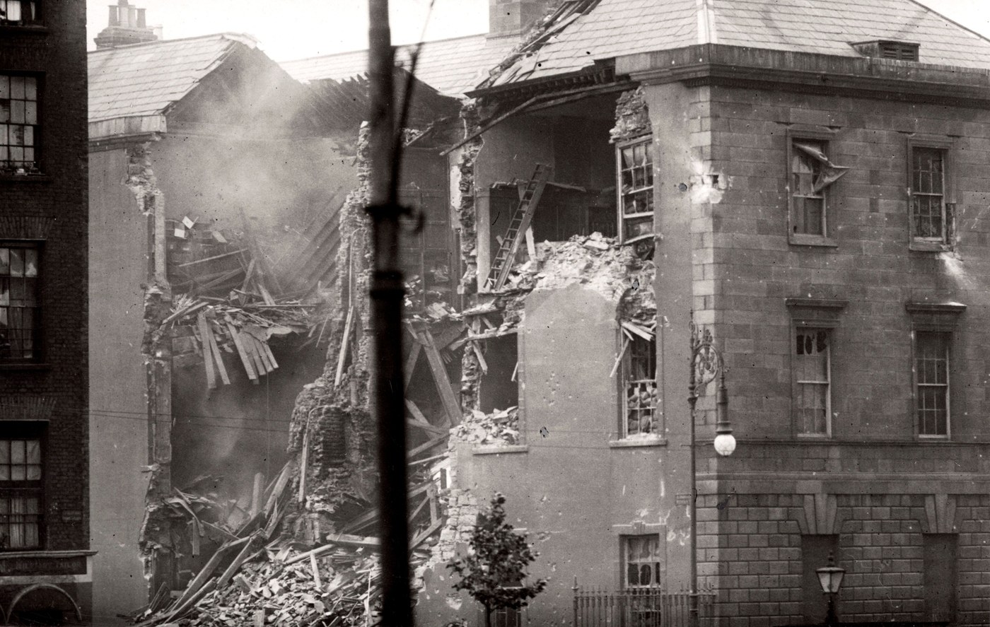Black and white photograph of a large stone building with a collapsed corner, rubble heaped in the street and a ladder against the broken wall.