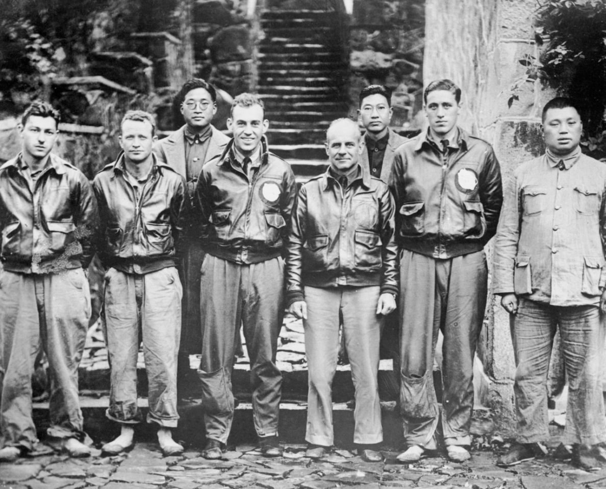 Black and white photograph of American airmen in flight jackets standing with Chinese men in front of steps