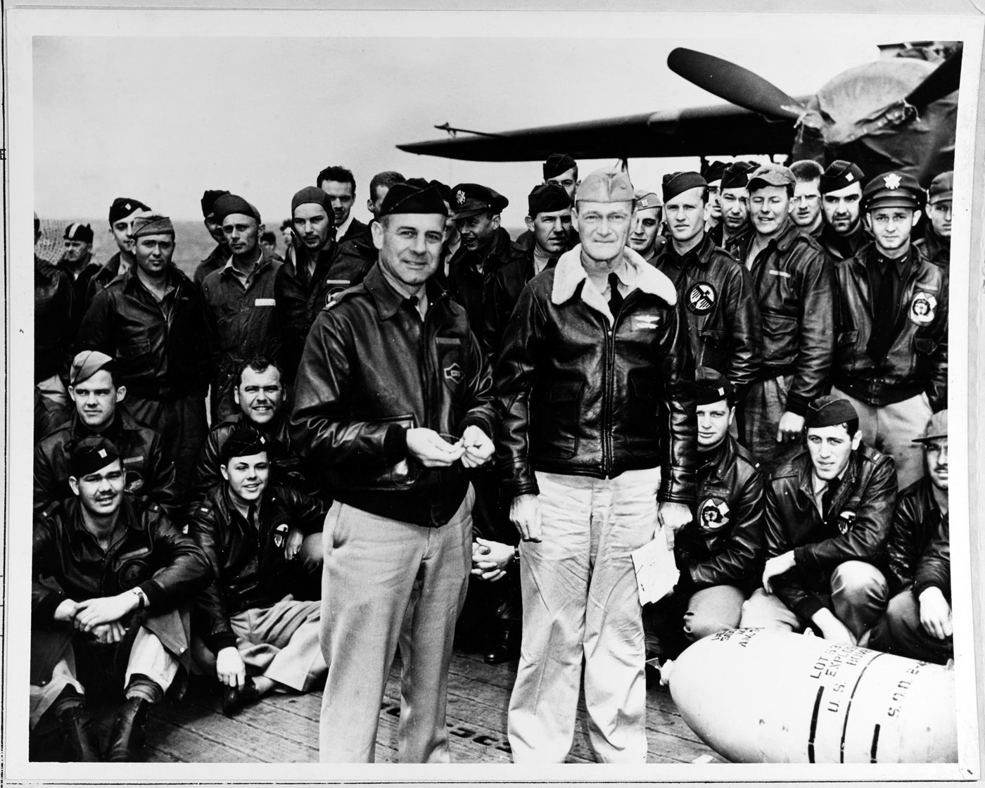 Black and white photograph of airmen and naval officers posing around a bomb on a carrier deck