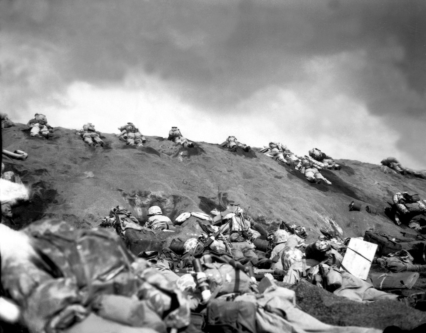 Black and white photograph of Marines lying prone on a slope of volcanic sand under drifting smoke
