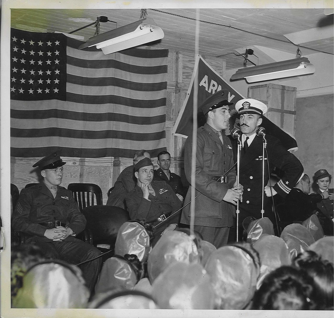 Black and white photograph of Basilone in uniform at a microphone beneath an American flag