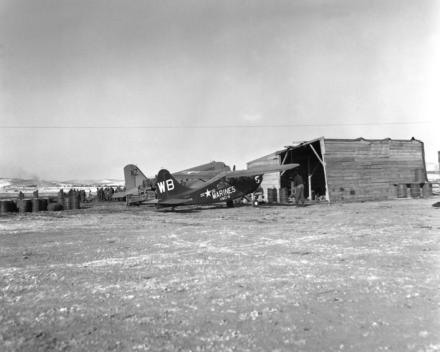 Black and white photograph of a small Marine observation plane marked WB and VMO-6 and a twin-engine transport parked on a frozen airstrip beside a wooden shed, with soldiers gathered in the background.